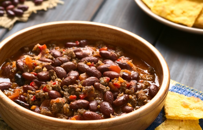 Wooden bowl of chili con carne with homemade tortilla chips in the back, photographed with natural light (Selective Focus, Focus in the middle of the chili)